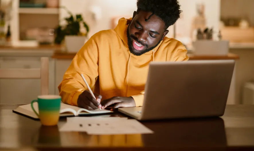 A person in a yellow hoodie sits at a table, writing in a notebook with an open laptop in front of them. A mug and some papers are also on the table. The setting appears to be a cozy, warmly lit home.