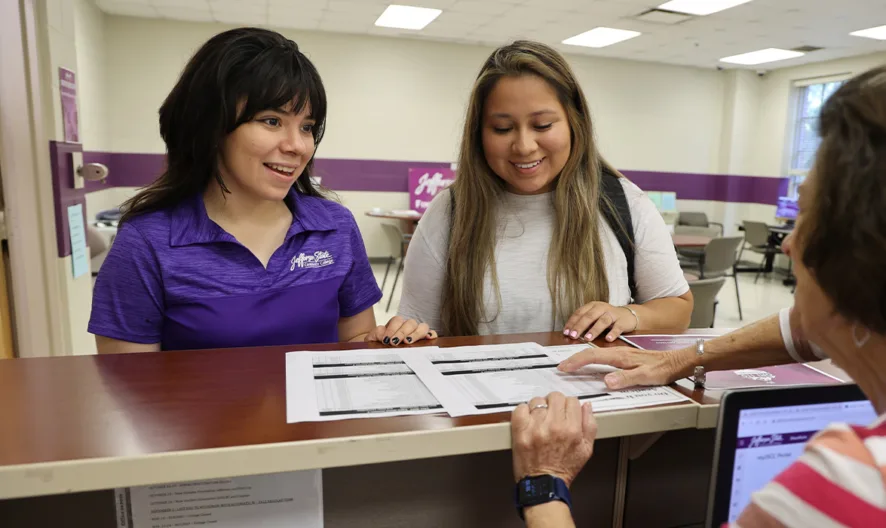 A woman behind a reception desk helps two women standing in front of her, showing them printed documents. The women are smiling in a bright, office-like setting with purple accents.