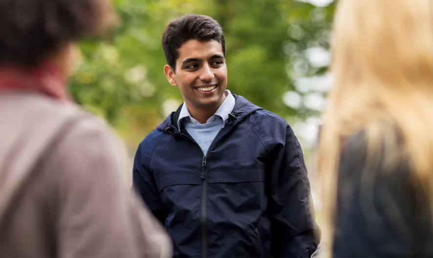 A young man in a navy jacket stands outdoors, smiling and talking with two other people whose backs face the camera, against a blurred background of green trees.