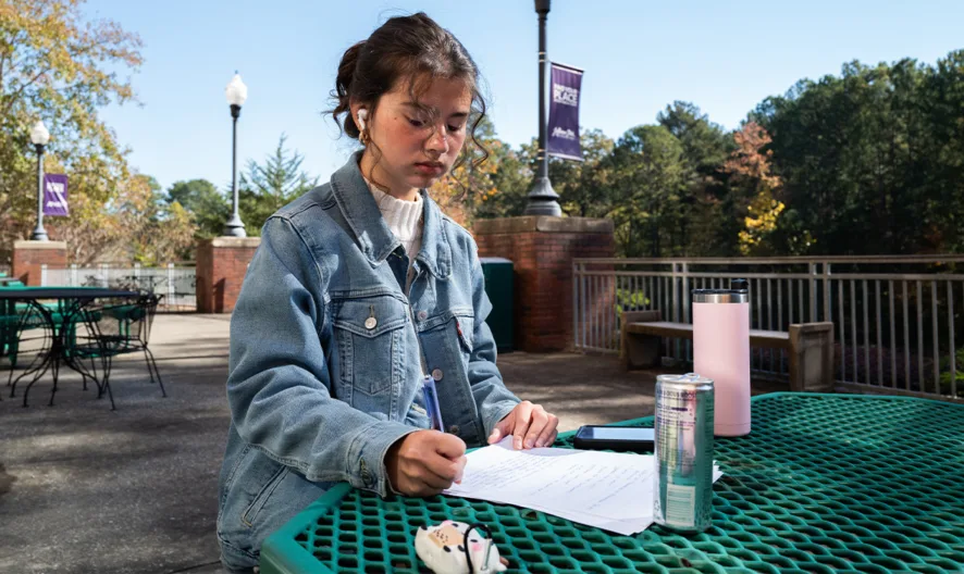 A young woman sits alone at a green outdoor table, studying papers and writing. She wears a denim jacket, with notebooks, a pencil case, and a pink bottle on the table. Trees and campus banners are in the background.