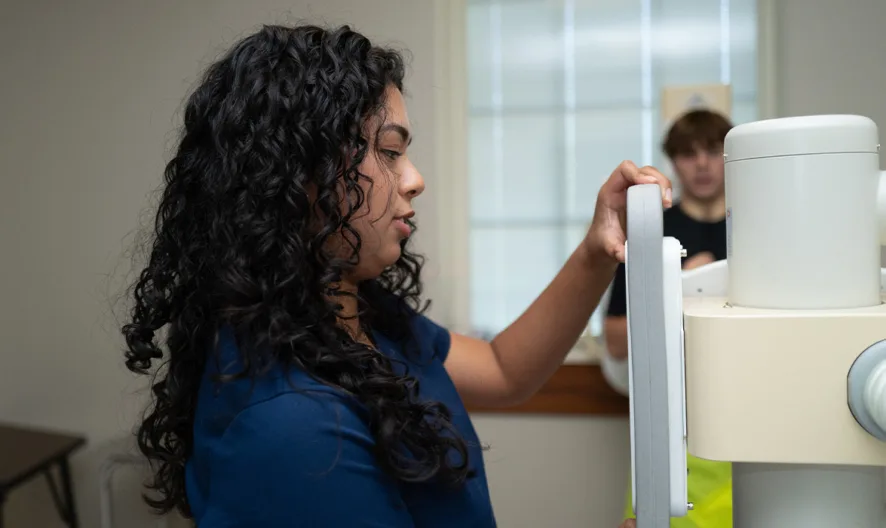 A woman with curly hair operates a medical imaging machine in a clinical room, while another person stands in the background near a window.