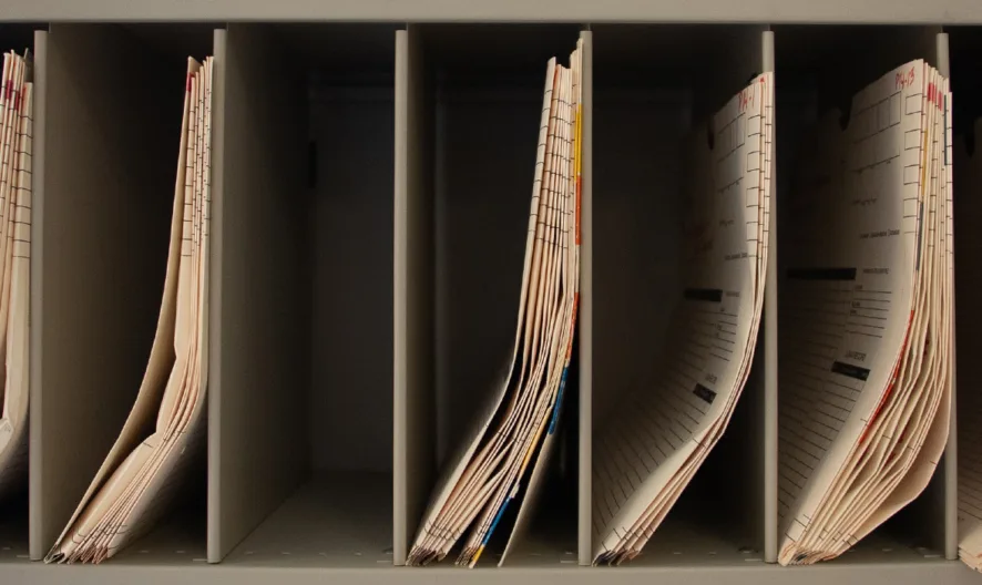 A close-up of a metal shelf with several vertical slots, each filled with organized manila file folders labeled with tabs.