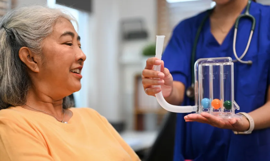 An older woman smiles while a healthcare professional in blue scrubs holds a breathing exercise device with colored balls in front of her, assisting her in a medical or rehabilitation setting.