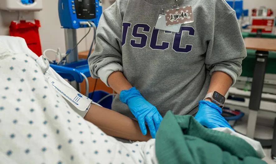 A healthcare worker in blue gloves and an ISCC sweatshirt tends to a patient in a hospital gown, holding the patients arm in a medical setting with equipment and supplies in the background.