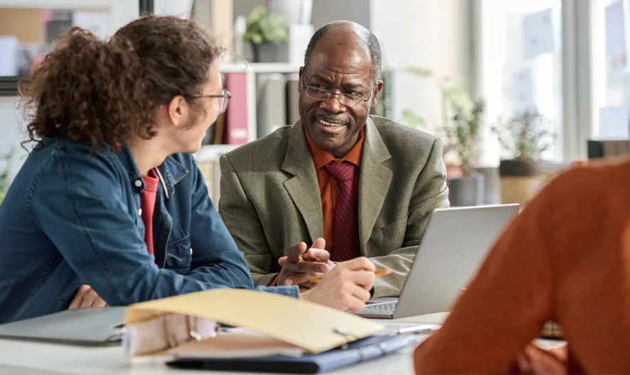 Three people sit at a desk in an office, engaged in conversation. One man in a suit smiles while speaking to a woman with curly hair; another person is seen from behind. Laptops and folders are on the table.