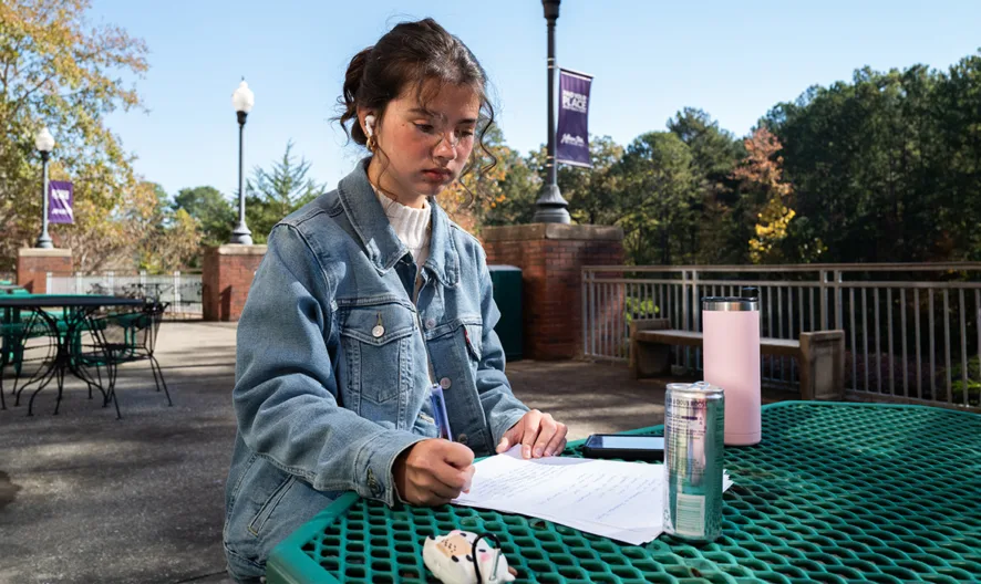 A person sits at a green metal outdoor table, writing on papers with a pen. They wear a denim jacket and have earbuds in. A pink water bottle, silver can, and pencil pouch are on the table. Trees and campus banners are in the background.