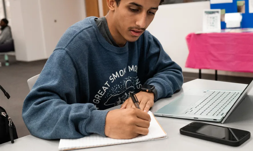Young person in a blue sweatshirt sits at a table, writing in a notebook next to an open laptop and a smartphone, with a backpack nearby in a modern indoor setting.