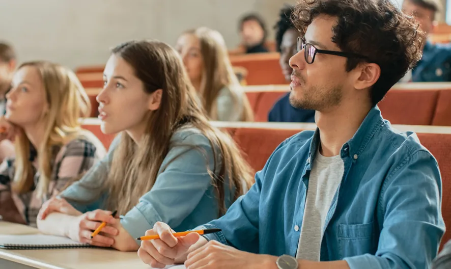 Students sit in rows in a classroom, attentively listening and taking notes. The atmosphere appears focused and engaged, with notebooks and pens visible on the desks.