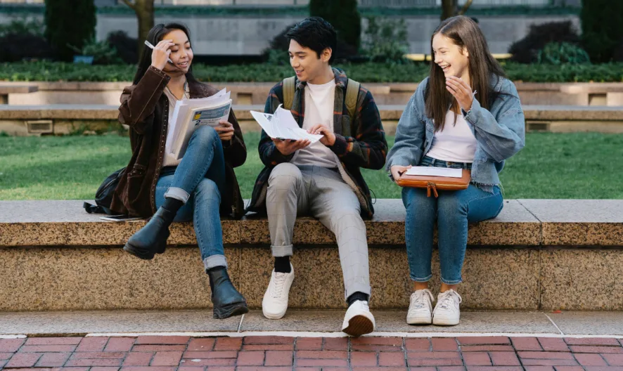 Three students sit on a stone bench outdoors, smiling and talking while holding notebooks and papers. They appear to be studying together on a college campus with grass and trees in the background.