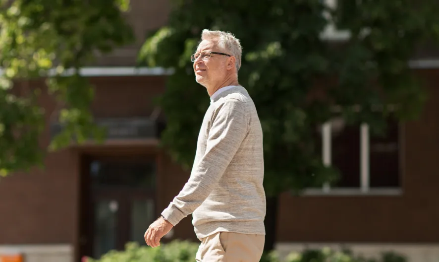 An older man with gray hair and glasses walks outdoors on a sunny day, wearing a light sweater and beige pants. Trees and a brick building are visible in the background.