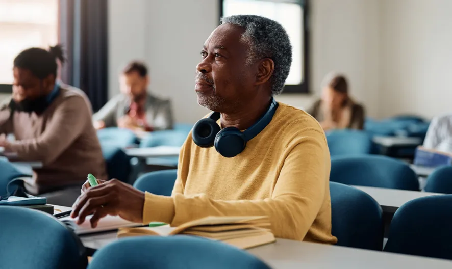 An older man wearing headphones around his neck sits in a classroom, taking notes and looking attentively ahead; other adults are seated behind him, focused on their work.