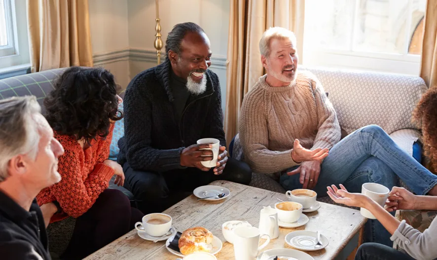 Five adults sit around a coffee table in a cozy living room, chatting and smiling while enjoying hot drinks and pastries. Sunlight streams in through the windows, creating a warm and inviting atmosphere.