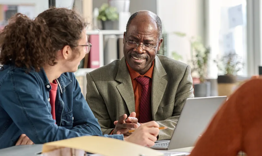 Three people sit around a table in an office, having a discussion. One man in a suit speaks while the others listen. There are papers, a laptop, and office supplies on the table.