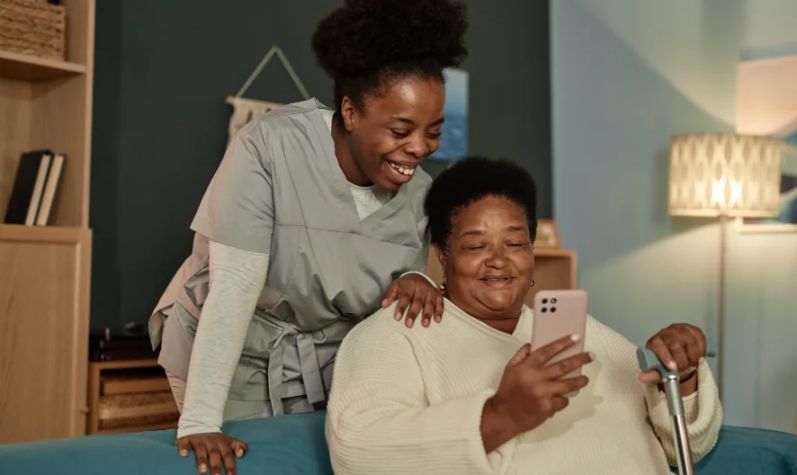 A caregiver in scrubs smiles and leans over the shoulder of an older woman sitting on a couch, who is holding a smartphone and a cane. Both appear happy and engaged with the phone.