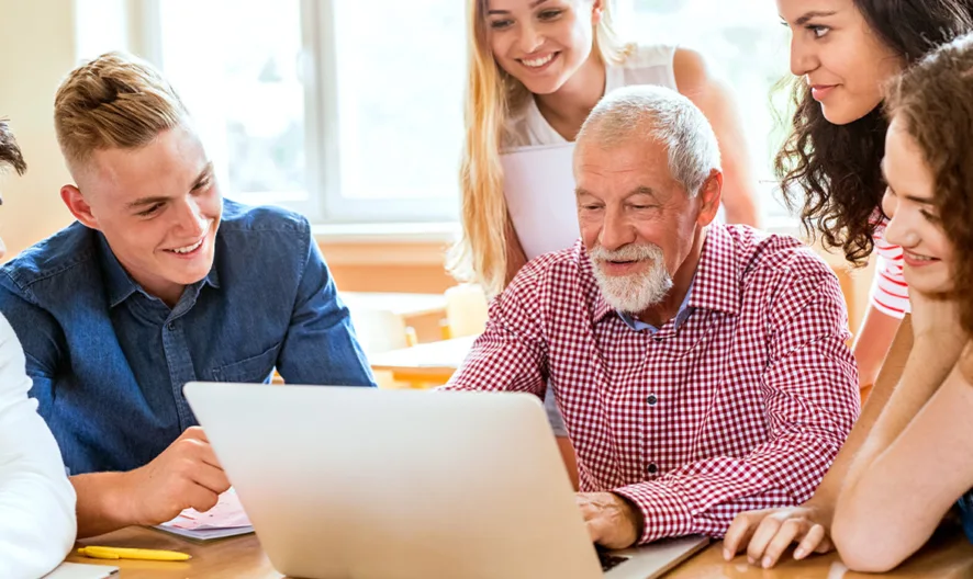A group of young adults and an older man with a beard, all smiling and gathered around a laptop. They appear to be having a discussion or working together in a bright room with large windows.