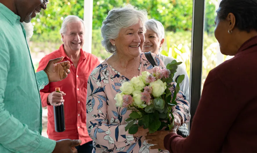 A smiling elderly woman holding a bouquet of flowers is greeted at the door by a friend, while two other smiling people stand behind her, one holding a wine bottle. Greenery is visible outside.