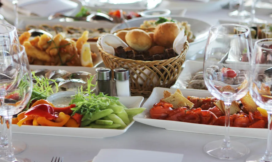 A table set for a meal with empty wine glasses, plates of assorted appetizers including vegetables and bread, and a basket of rolls, all arranged on a white tablecloth.