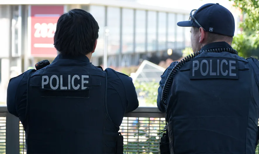 Two police officers in uniform stand outdoors with their backs to the camera, facing a railing. Trees and a building are visible in the background on a sunny day.