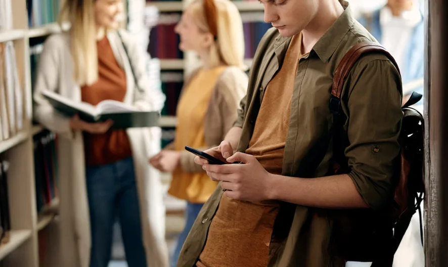 A student with a backpack leans against a bookshelf in a library, looking at a smartphone. In the background, two people stand and talk, one holding an open book. Shelves filled with books line the scene.