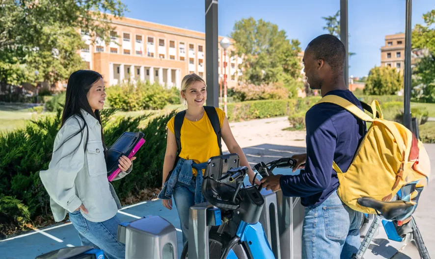 Three students gathered around a bike rack having a conversation, they are smiling.