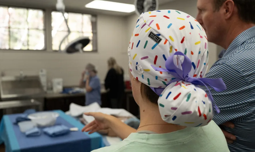 A person wearing a white cap with colorful sprinkles and a purple bow stands beside a man in a striped shirt, both looking at a medical setting with people and supplies in the background.