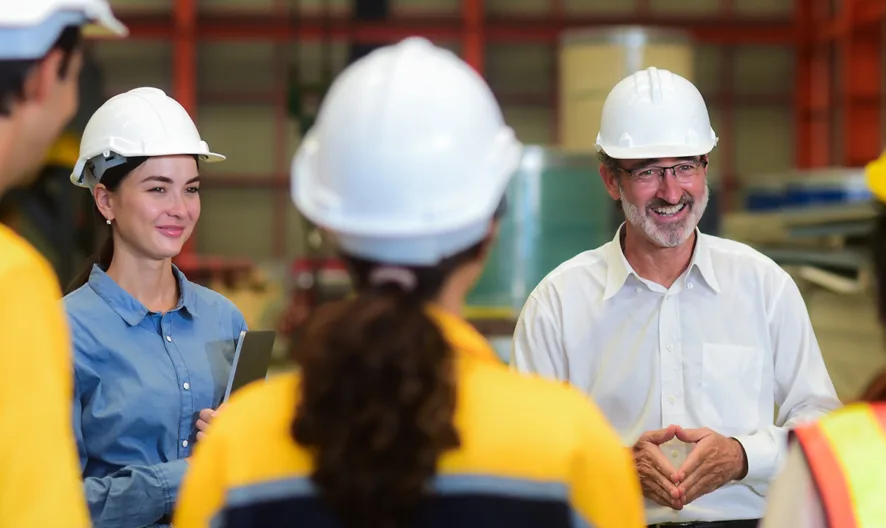 A group of workers wearing safety helmets and high-visibility clothing listen to a man in a white shirt and hard hat, standing and smiling inside an industrial warehouse or factory setting.