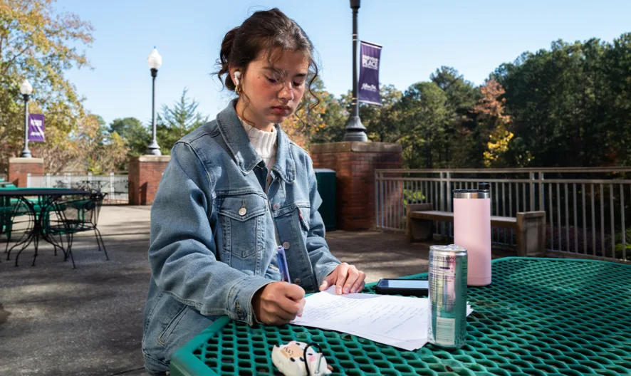 A young person sits alone at a green outdoor table, writing on papers. They wear a light denim jacket and have a drink bottle nearby. Trees and campus banners are visible in the background.