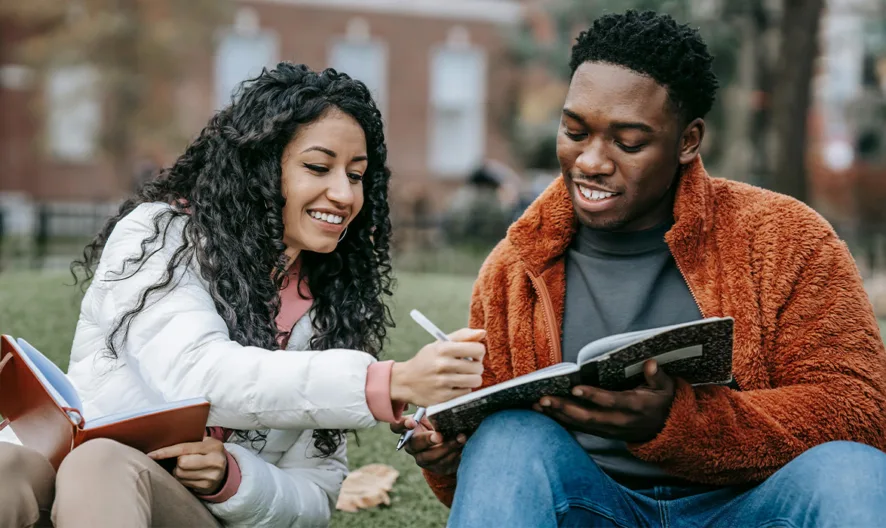 Two college students sit on grass outside, smiling and studying together. One writes in a notebook while the other holds it open. Books and autumn leaves are visible around them.