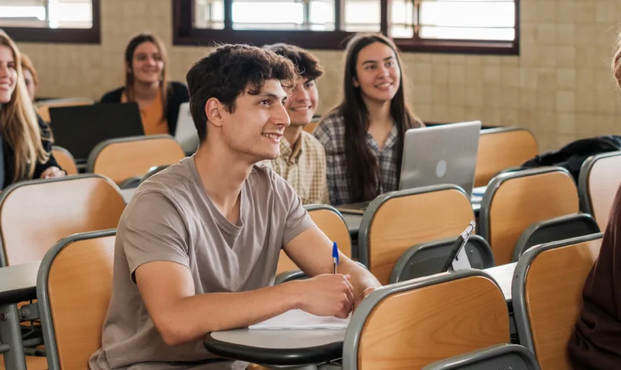 A group of students sit at desks in a classroom, attentively listening and smiling. Some have laptops, others write in notebooks. Sunlight streams through windows in the background.
