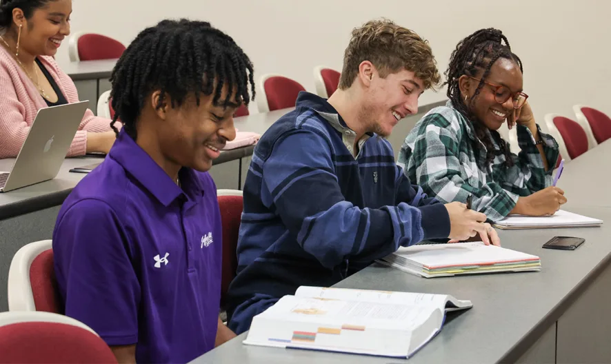 Three students sit at desks in a classroom, smiling and writing in notebooks, while a fourth student sits behind them working on a laptop. The room has rows of red and white chairs and gray desks.