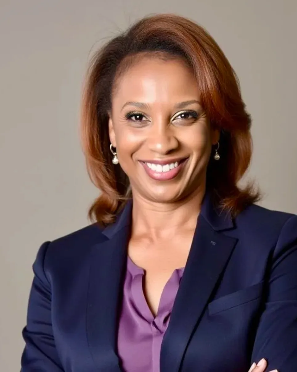 A woman with shoulder-length brown hair smiles confidently. She is wearing a navy blazer over a purple blouse, with small hoop earrings, standing against a plain light background.