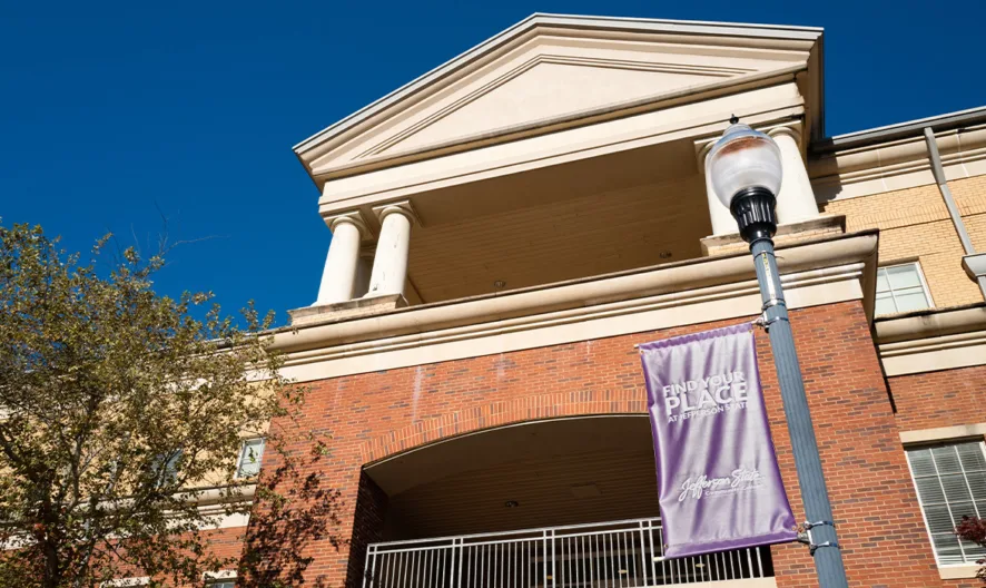 A red and tan brick building with white columns and pediment is shown under a clear blue sky. A purple banner reading “The Place” hangs from a lamp post in front, and tree branches are visible on the left.
