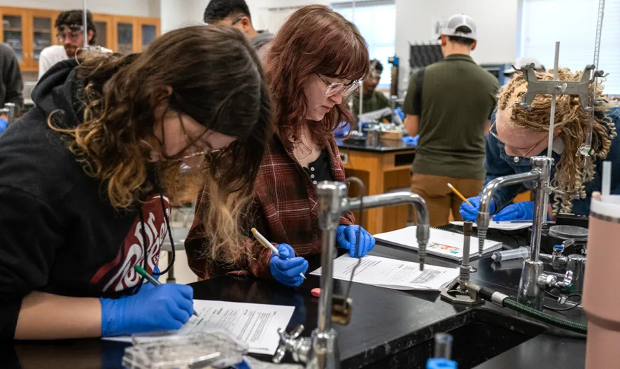Students wearing blue gloves work together on science experiments and fill out worksheets in a busy laboratory classroom with lab equipment, sinks, and wooden cabinets visible in the background.