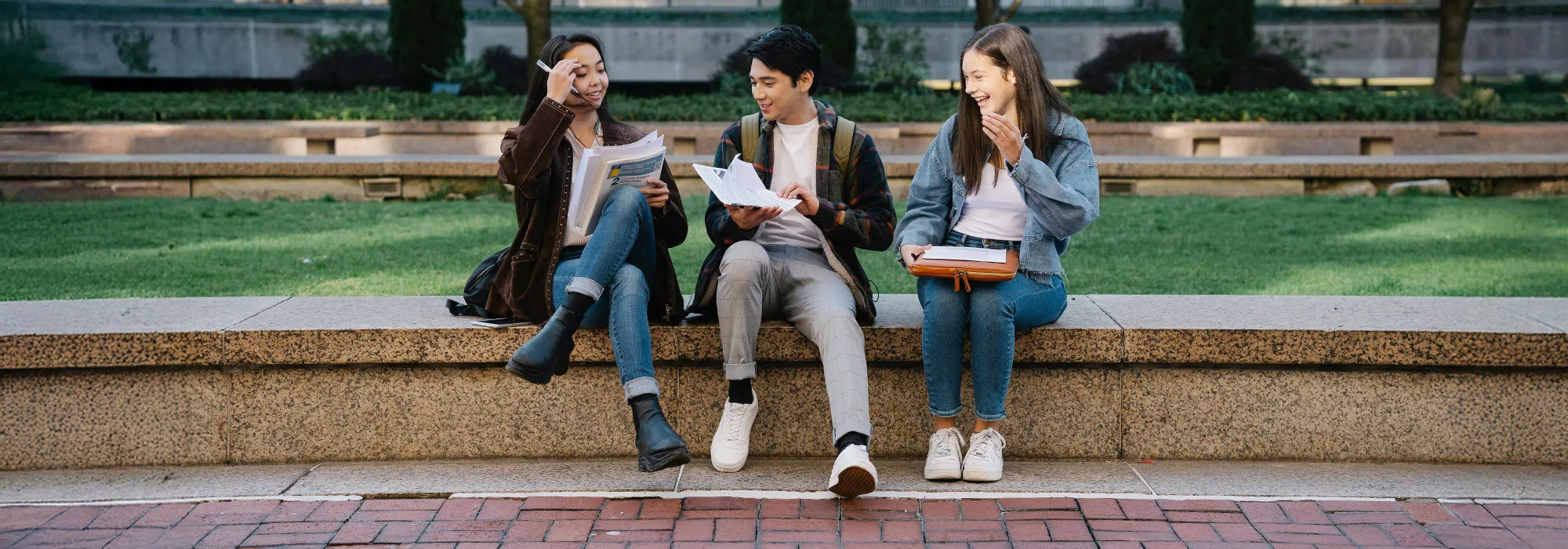 Three students sit on a low stone wall outdoors, smiling and talking while holding books and notebooks. Grass and trees are in the background. It appears to be a university or college campus.