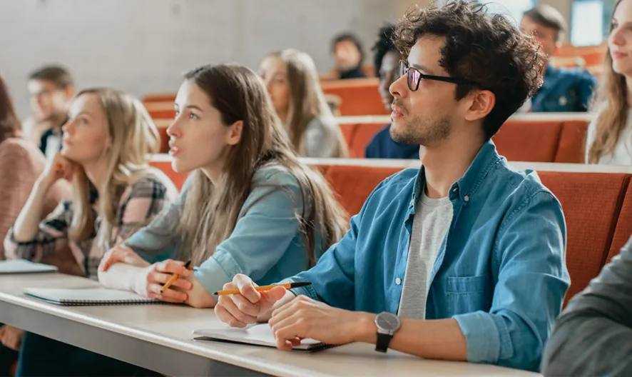 Students sit in rows in a lecture hall, attentively listening and taking notes. The classroom is bright, and the students appear focused and engaged in the lesson.