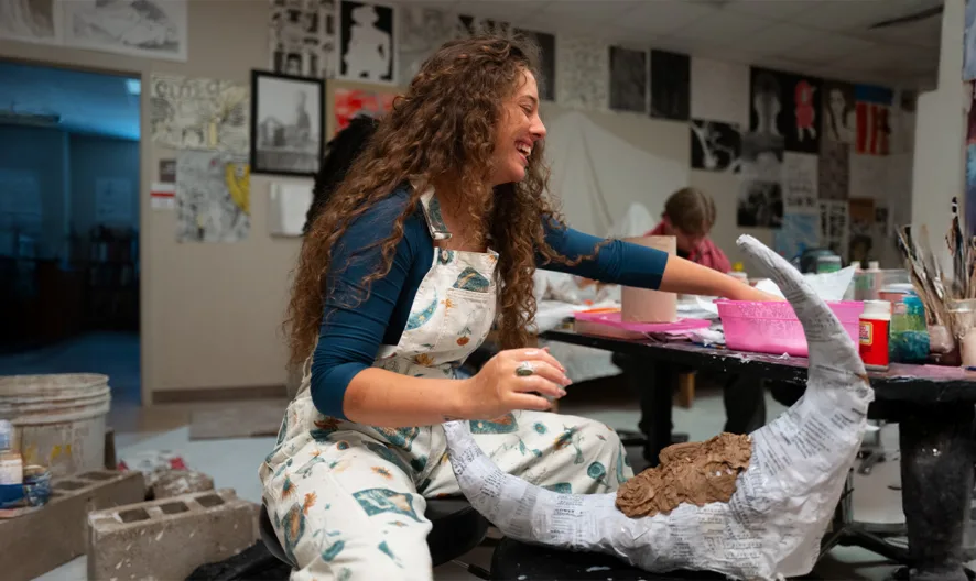 A woman in paint-splattered overalls smiles while working on a large horned sculpture in an art studio filled with paintings, sketches, and art supplies.