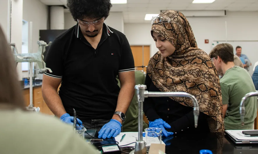 Students in a science lab work together on an experiment. A man wearing gloves stands next to a woman in a patterned hijab, both focused on a task. Other students work in the background.