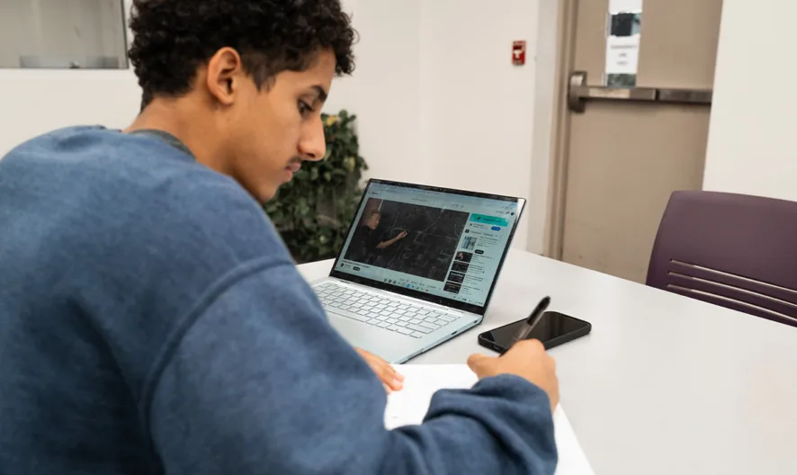 A young person with curly hair sits at a table, writing in a notebook, with an open laptop and smartphone in front of them. The setting appears to be a modern, bright study or office space.