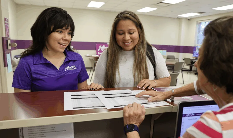 Two young women smile while talking to an older woman at a reception desk. The older woman points to papers on the counter, and the setting appears to be an office or student service center with purple accents.