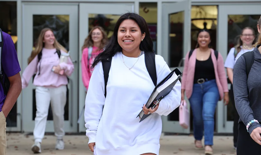 A group of smiling students, carrying backpacks and notebooks, walk out of a school building together. The students appear happy and relaxed as they head outside.