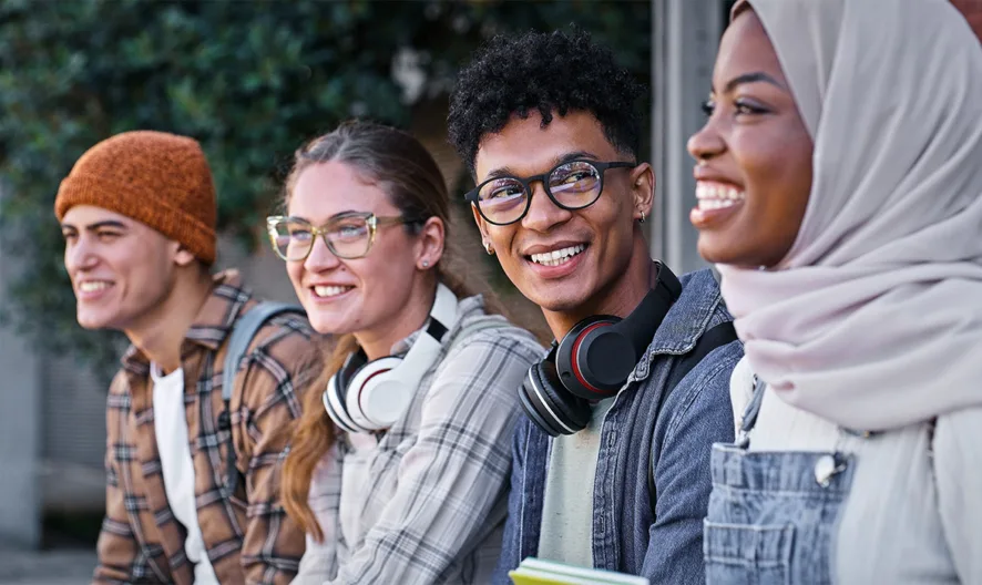 Four young adults sit outdoors, smiling and talking. They wear casual clothes, two have headphones around their necks, and one wears a headscarf. They appear to be friends or students enjoying each other’s company.