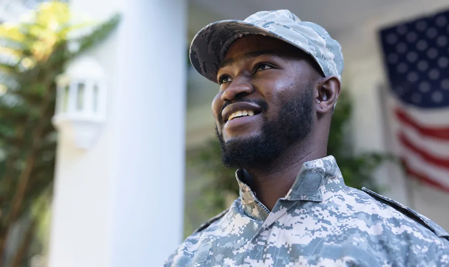 A man in military camouflage uniform and cap smiles while standing outdoors, with a blurred American flag hanging in the background. Lush greenery and a white building are also visible.