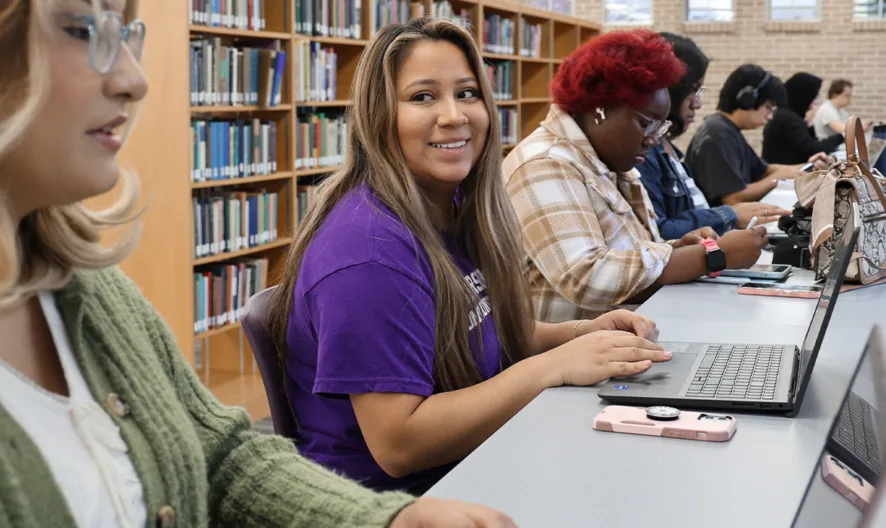 Several students sit at a long desk in a library, working on laptops. One student in a purple shirt smiles at the camera while others focus on their screens or phones. Bookshelves line the background.