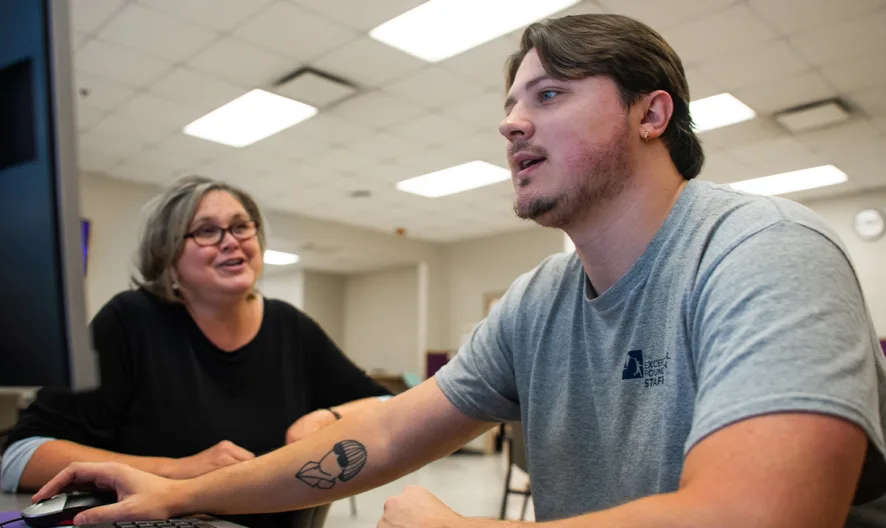 A young man in a gray shirt uses a computer while a woman with glasses sits beside him, smiling. They are in a classroom or office with fluorescent lighting and a clock on the back wall.