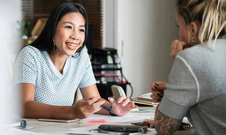 Two women sit across from each other at a desk, engaged in conversation. One woman is smiling and holding a pen, while the other listens attentively. Papers, notebooks, and a phone are spread out on the desk.