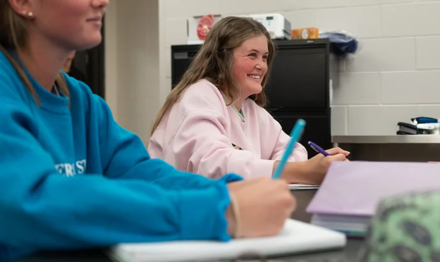 Two students sit at desks in a classroom, smiling and writing in notebooks. Classroom supplies and a cabinet are visible in the background.