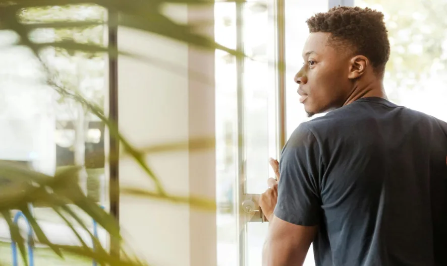 A young person wearing a dark t-shirt and carrying a backpack looks over their shoulder while exiting through a glass door, with greenery visible both inside and outside.