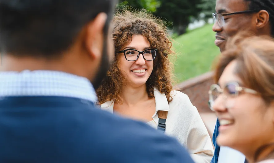 A group of four people stands outside, smiling and talking. Three people are facing each other, while one person with curly hair and glasses looks at them, outdoors on a sunny day.