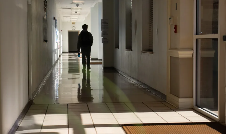 A person with a backpack walks down a brightly lit hallway, casting a shadow on the shiny floor. The corridor is lined with doors and windows, with natural light streaming in through a glass entrance.