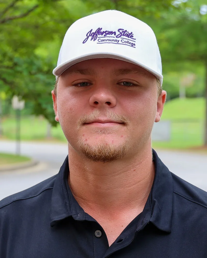 A young man wearing a white Jefferson State Community College cap and a black collared shirt stands outdoors, with greenery and a road blurred in the background.